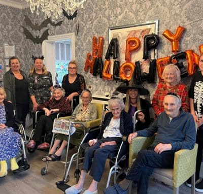 Group of people in costumes celebrating Halloween in a decorated room with 'happy Halloween' balloons in the background.